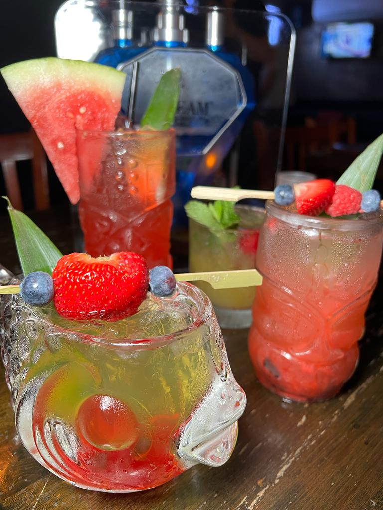 A variety of colorful cocktails on a bar counter. Includes a watermelon-garnished drink, a fruity cocktail in a fish-shaped glass, and a tropical drink with berries and a pineapple slice.