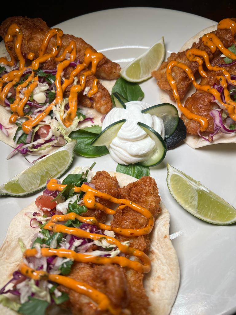Three fish tacos on a white plate with lime wedges, shredded cabbage, and a dollop of sour cream. The fish is battered and fried, and the tacos are drizzled with a spicy sauce.