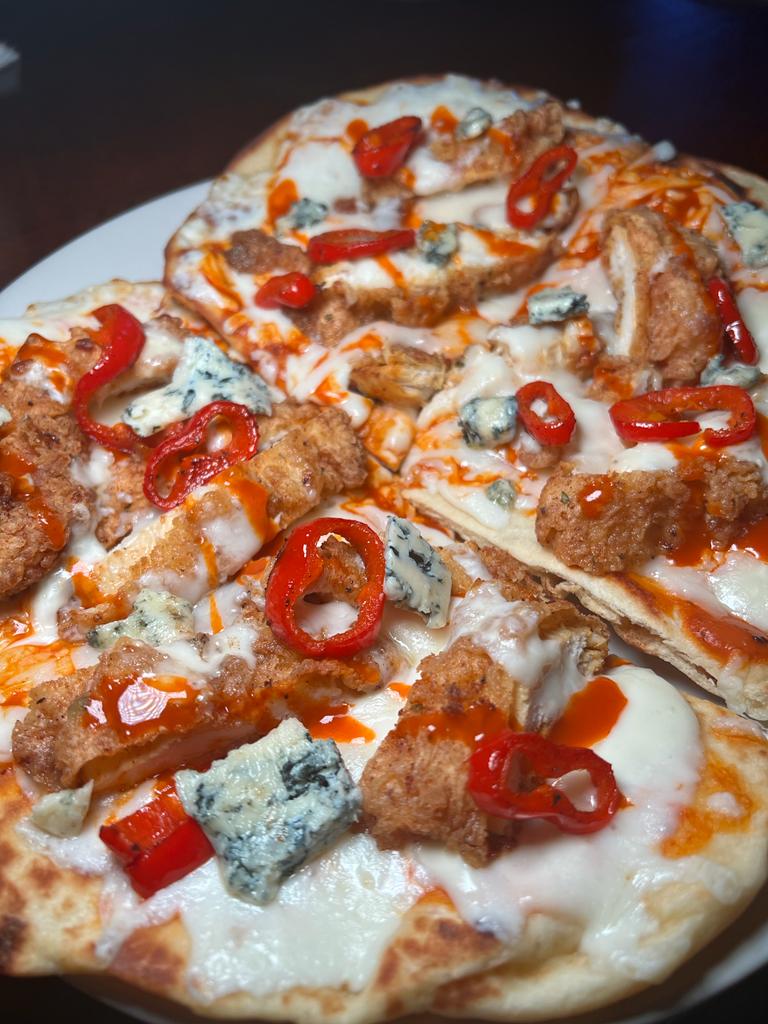 A close-up of a pizza with crispy chicken, blue cheese crumbles, red pepper flakes, and a drizzle of sauce.