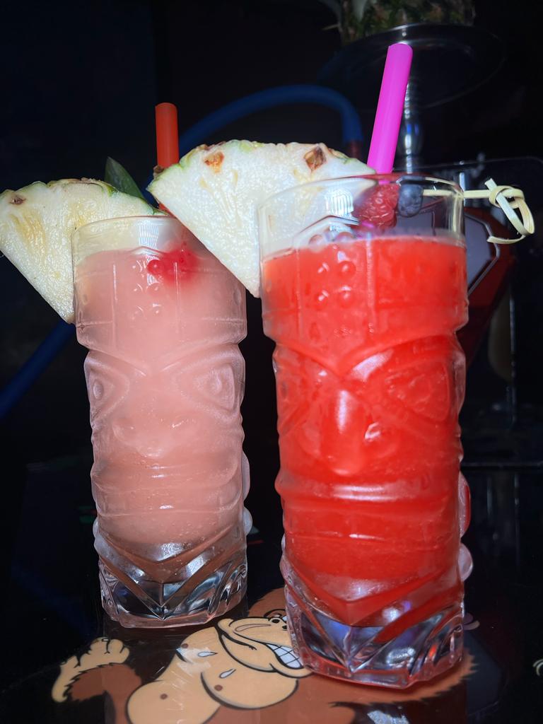 Two tiki-style glasses with colorful drinks and fruit garnishes on a cartoon-themed table. Hookah in the background.