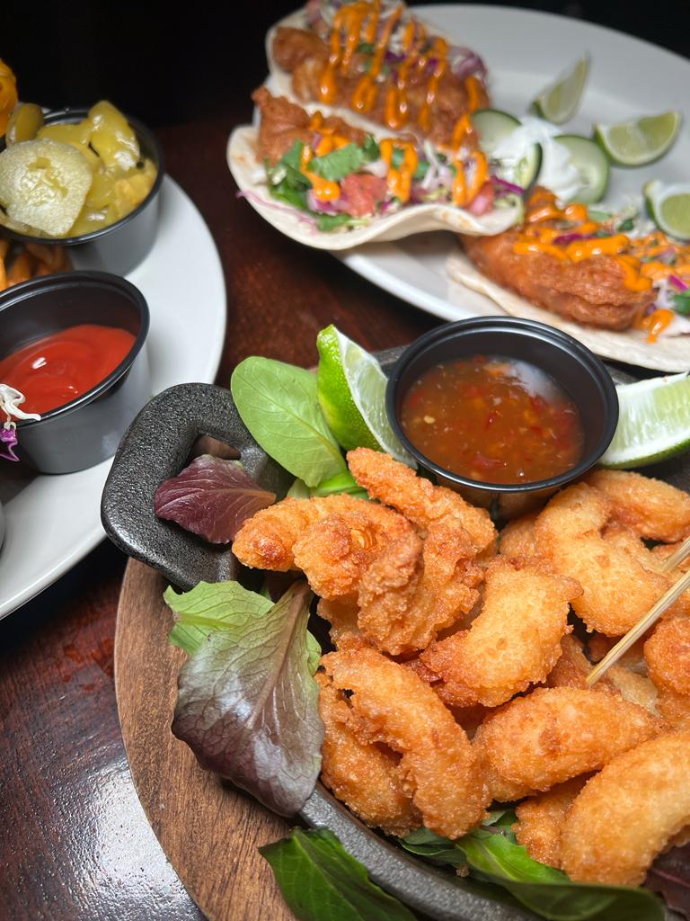 A close-up of a plate of fried shrimp with a side of dipping sauce and lime wedges. In the background, there are fish tacos and other appetizers.