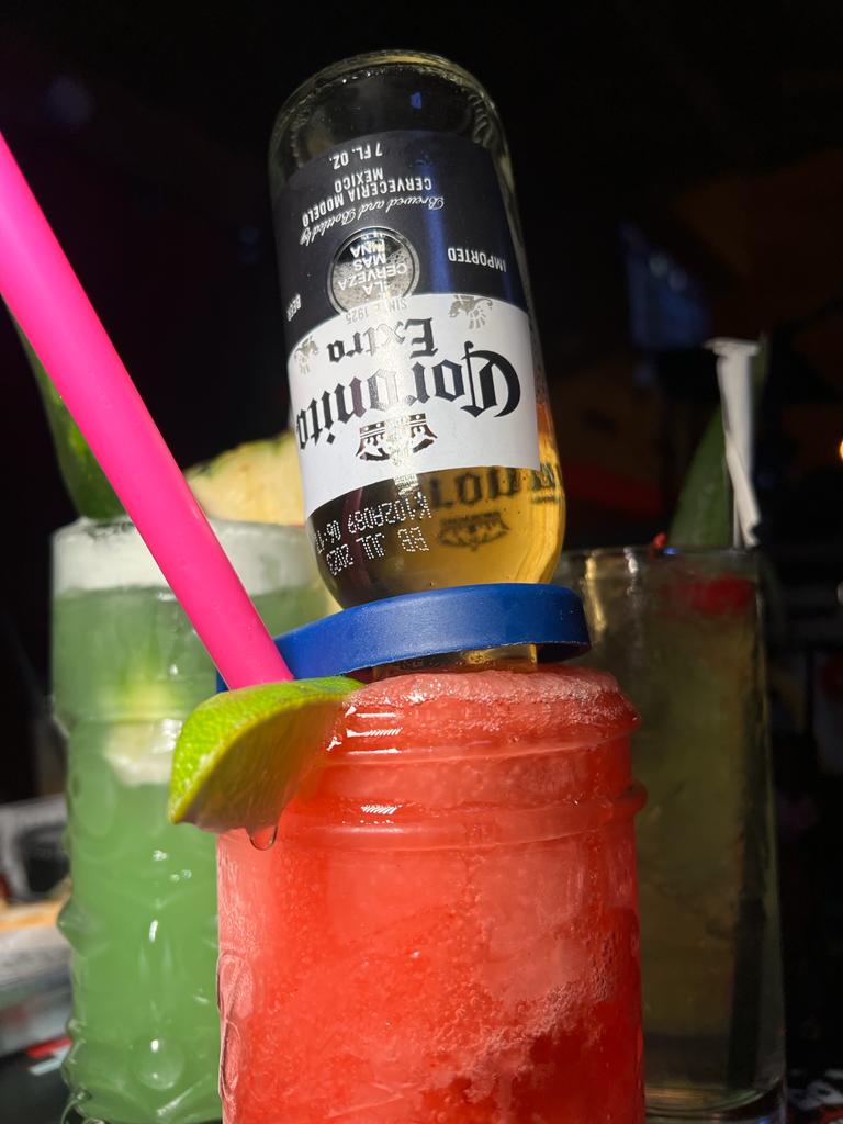 A Corona beer bottle chilling in a frozen margarita cocktail with a pink straw and a lime wedge.