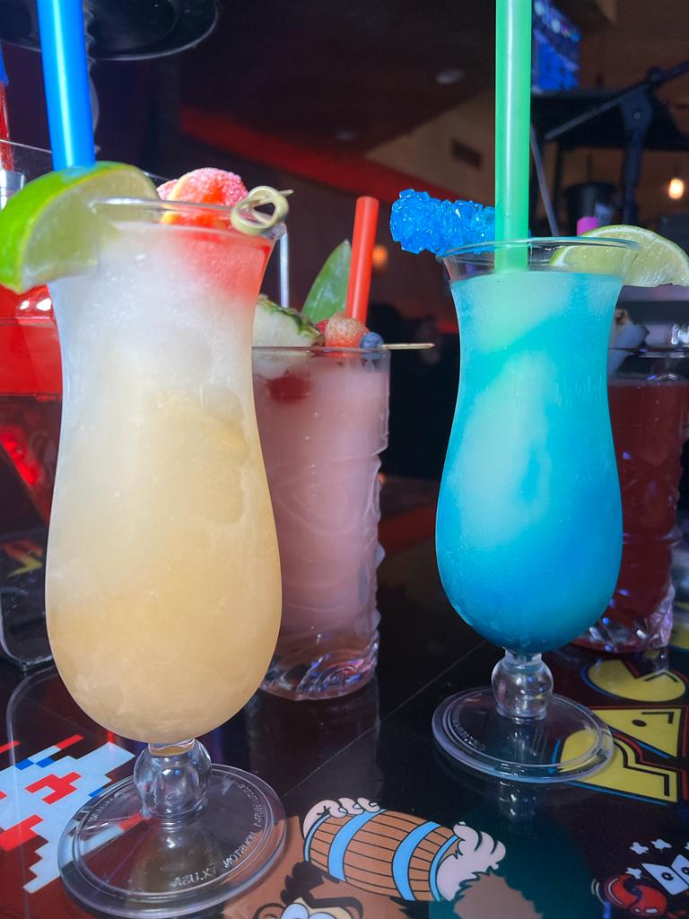 Three colorful frozen cocktails on a bar counter with a Pac-Man game mat underneath. The drinks include a yellow one, a pink one, and a blue one.