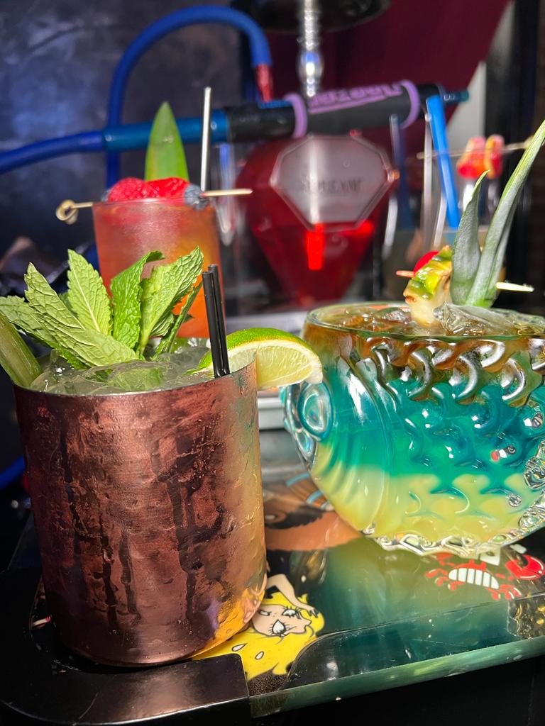Three colorful cocktails on a tray. One is a Moscow Mule in a copper mug, garnished with mint. The other two are in decorative glasses with pineapple and other tropical garnishes.
