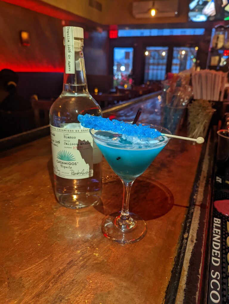 A vibrant blue margarita cocktail garnished with a blue rock candy stick, served in a martini glass on a bar counter. A bottle of Casamigos Blanco Tequila is visible in the background.