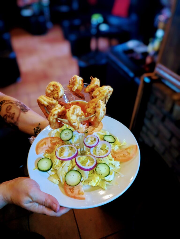 A plate of shrimp cocktail with a side salad. The shrimp are arranged in a martini glass and served with a dipping sauce. The salad includes lettuce, tomatoes, cucumbers, and onions.