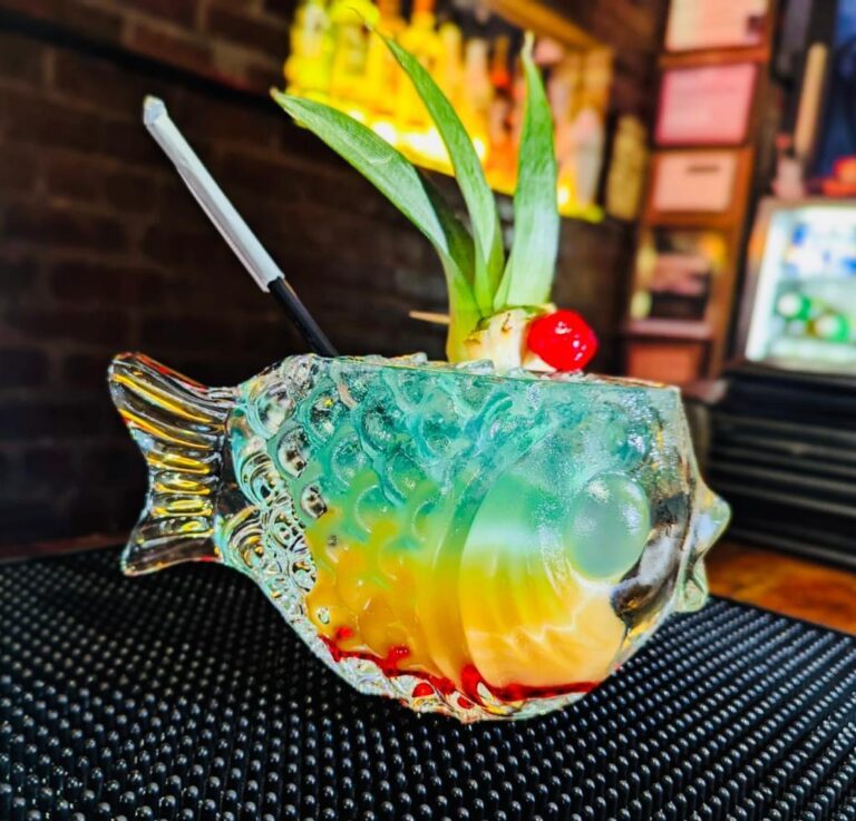 A colorful layered cocktail served in a fish-shaped glass. The drink has blue, yellow, and orange layers and is garnished with a pineapple slice and a cherry
